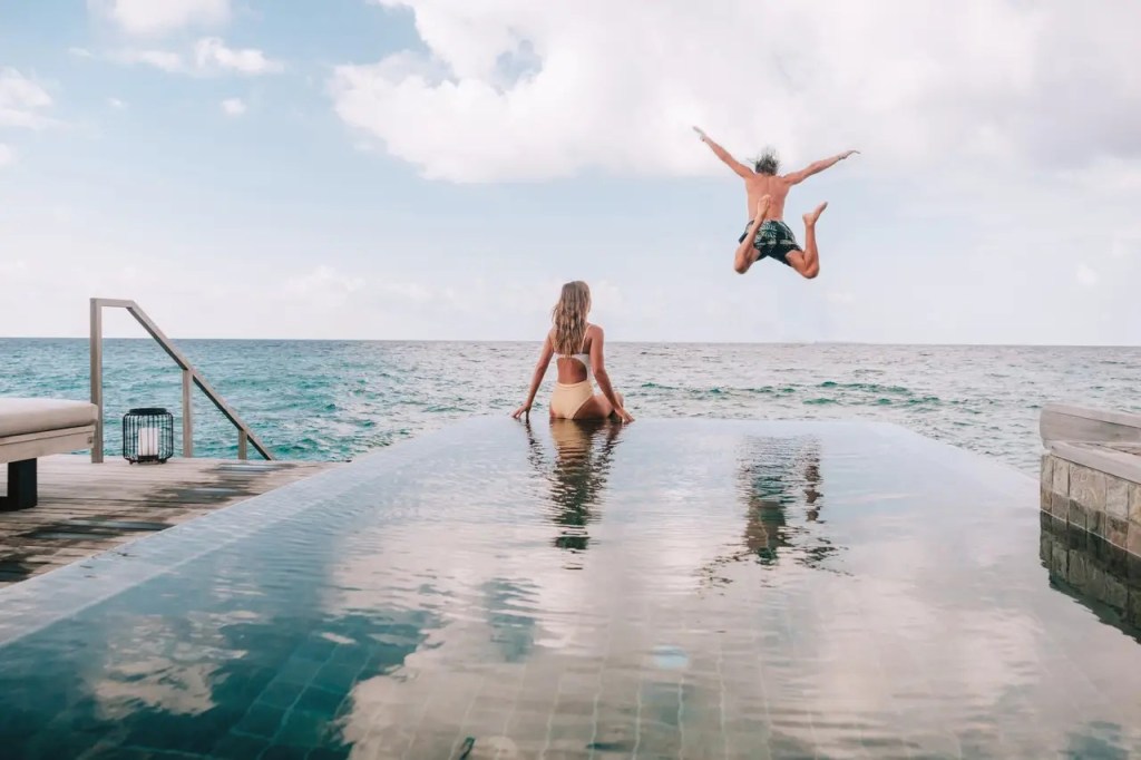 A woman sitting on the edge of a pool facing the ocean while a man jumps into the ocean